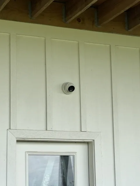 Small wireless security camera mounted on cream board and batten siding above a window.