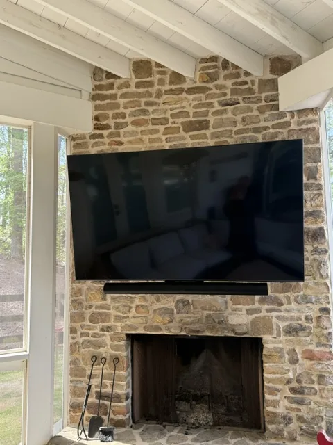 Wall mounted television and soundbar on a rough stone chimney under an exposed timber beam ceiling.