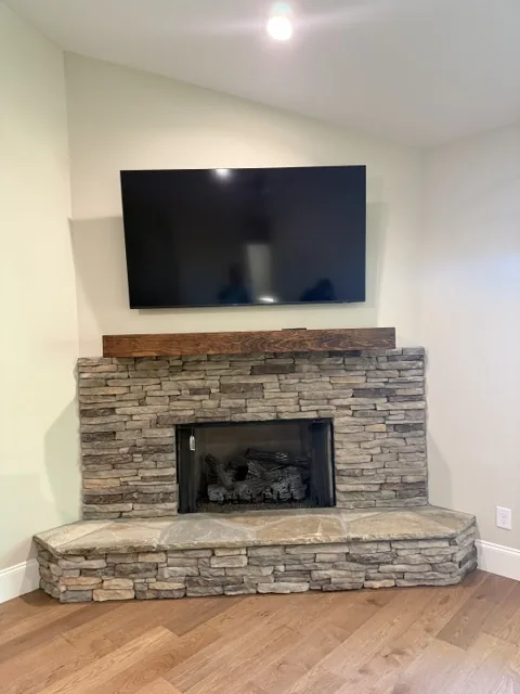 Television mounted above a stacked stone corner fireplace with a wooden mantel and hardwood floor.